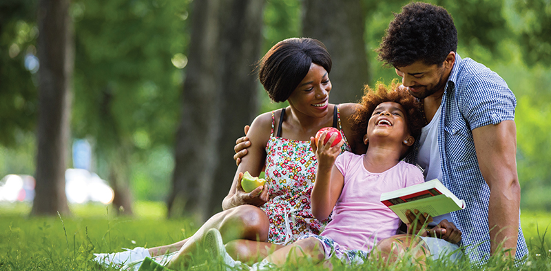 Mom, Dad and daughter are sitting in a park reading and eating apples